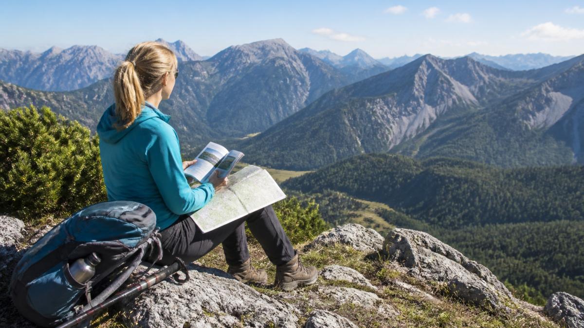 Wanderer sitzt auf sonnigem Berggipfel mit aufgeschlagenem Wanderbuch und Wanderkarte auf den Knien, blickt auf die Berglandschaft, klarer Himmel