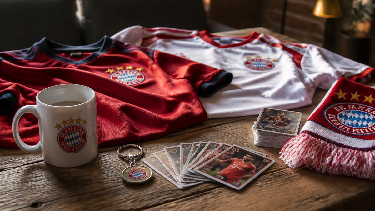Sammlung von Fußball-Fanartikeln auf einem Tisch arrangiert: Heimtrikot und Auswärtstrikot nebeneinander, weiße Tasse mit Vereinslogo, Schlüsselanhänger und Sammelkarten in warmer Beleuchtung
